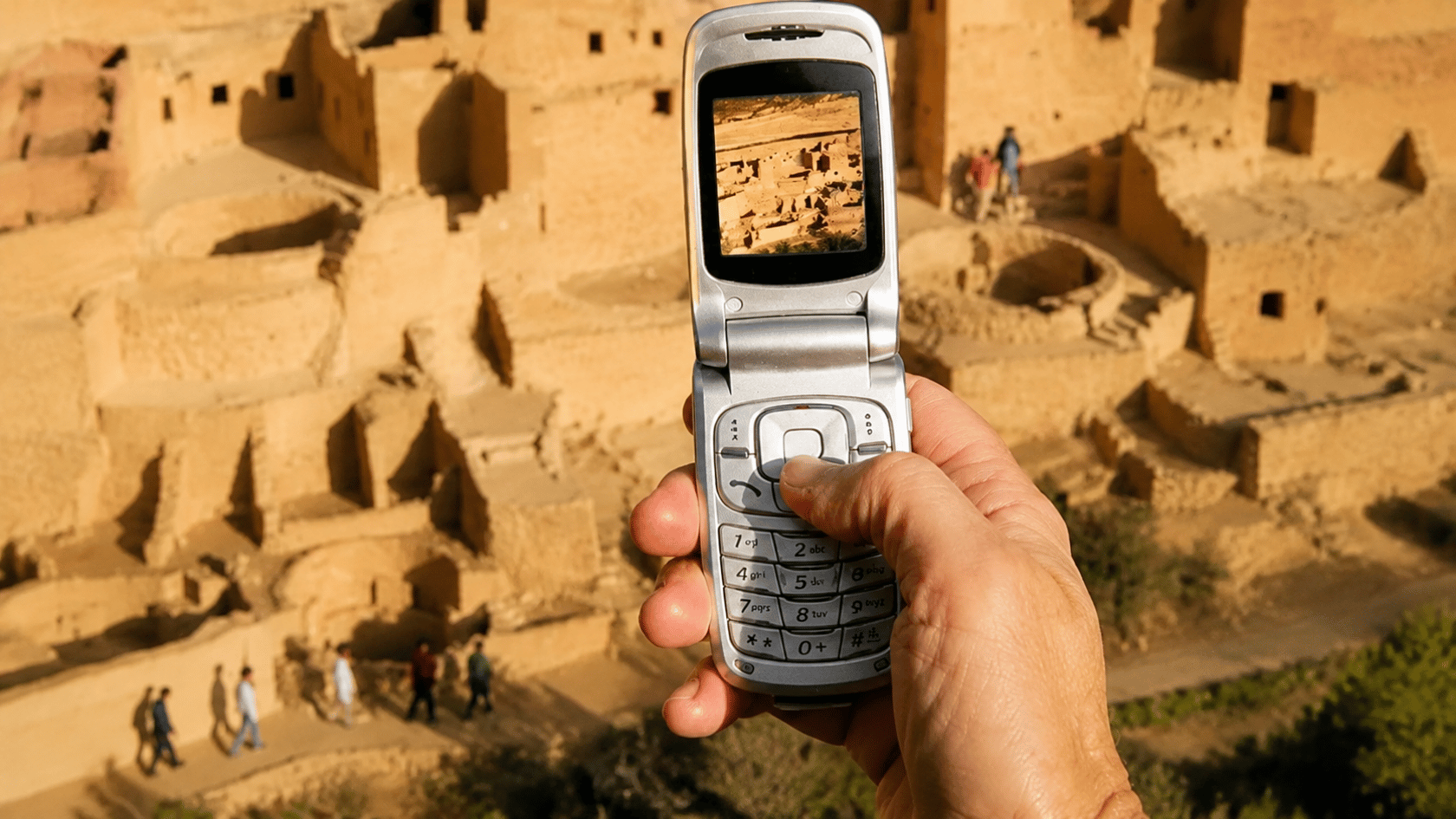 Hand holding a flip phone capturing ancient stone ruins, with the phone screen showing the same scene in the background.