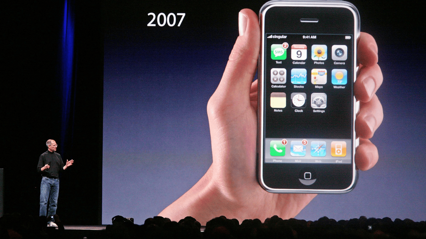 Steve Jobs presenting an early iPhone on stage with “2007” displayed on a large screen and an audience in the foreground.
