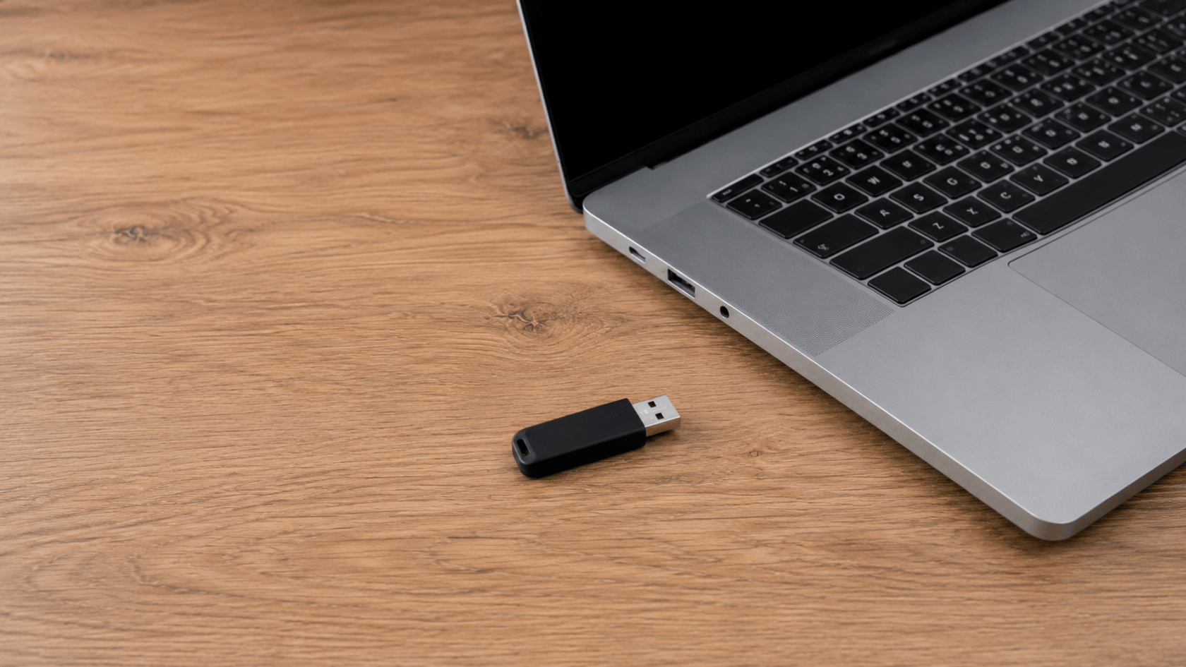 Laptop and USB flash drive on a wooden desk in a clean workspace, viewed from a slight top-down angle with neutral lighting.
