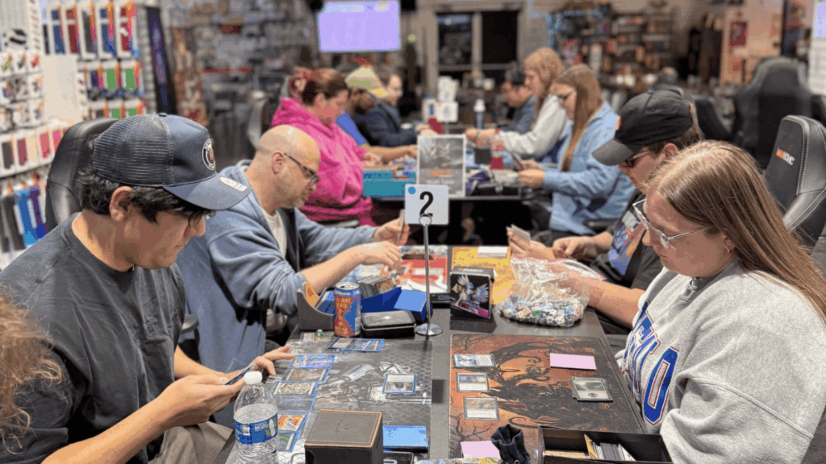 People seated at long tables in a game store playing trading card games with cards, deck boxes